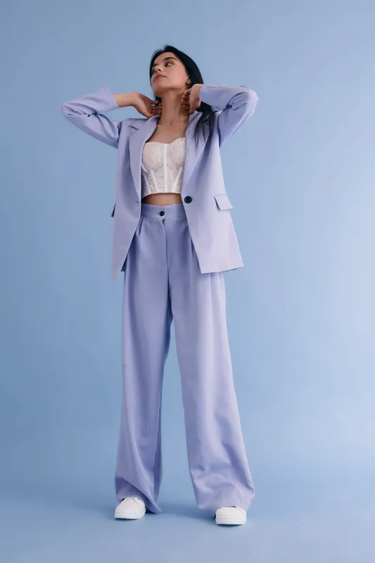 Woman in lilac suit poses confidently against pastel blue background, wearing white lace top and casual white shoes, showcasing modern fashion style.