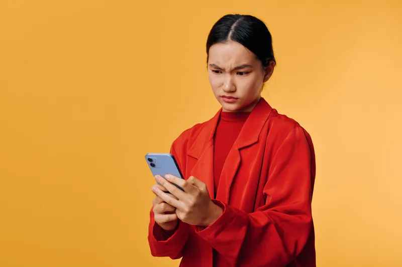 Woman in red jacket looking confused and concerned while holding smartphone with both hands on yellow background. Female user checking phone with serious expression in casual outfit.