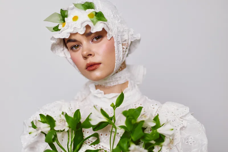 Elegant young woman wearing a vintage white dress with floral motifs, holding green plants and flowers, evoking themes of nature and femininity