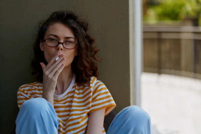 Casual woman with glasses sits against a wall in a sunny home, smoking a cigarette. Striped shirt and blue pants convey a relaxed lifestyle and intimate everyday moment.