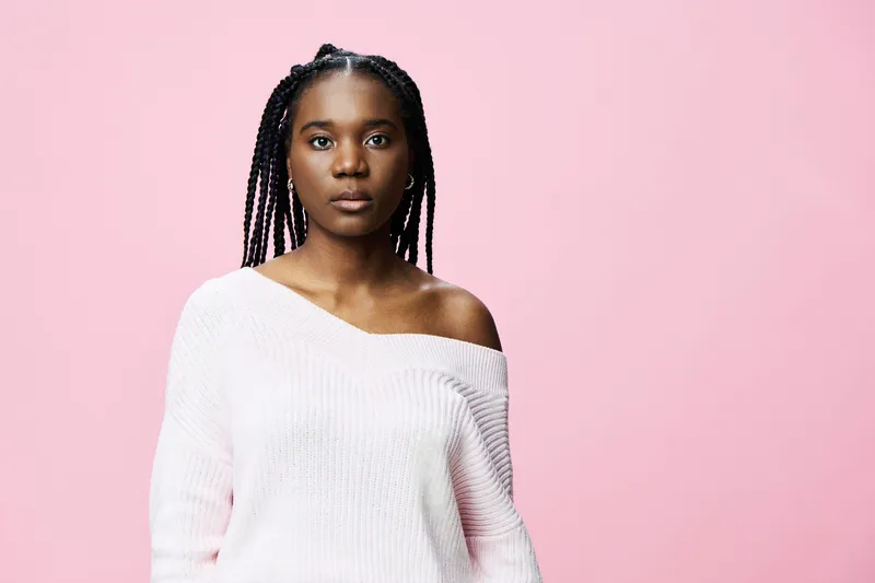 Confident young woman with braided hair in a cozy off shoulder sweater against a soft pink background, expressing empowerment and self love