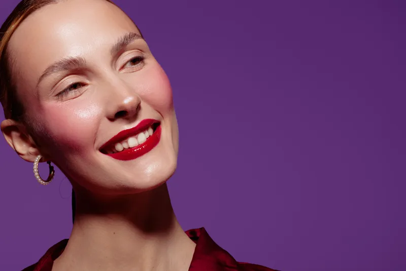 Close-up of a smiling woman with red lipstick and hoop earrings against a purple background. Concept of beauty, makeup, fashion, and confident female portrait in studio lighting.