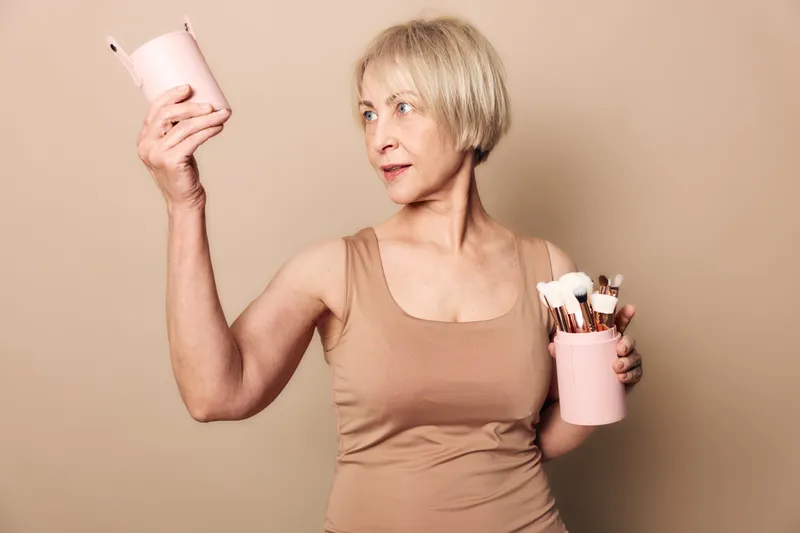 Mature woman with short blonde hair holds two pink containers with makeup brushes, looking aside. Studio portrait on beige background, concept for beauty tools and skincare routine.