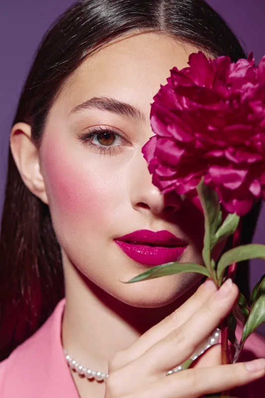 Young woman with vibrant magenta lipstick and blush holds bright red peony close to her face in a creative beauty and fashion concept on pink background.