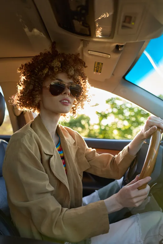 Woman driver sits in a retro car, wearing sunglasses and a beige coat, curly hair framed by sunlight, portraying a confident lifestyle moment with warm film color