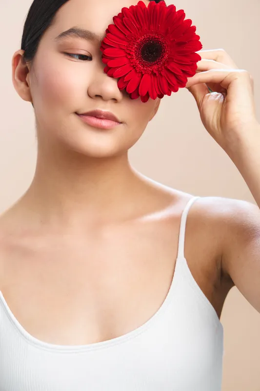 asian woman holds a red flower near her eye in a soft studio setting, conveying calm elegance and natural beauty with a gentle expression and minimal wardrobe.