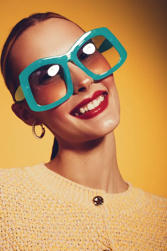 Smiling woman with large turquoise sunglasses and red lipstick poses against yellow background. Fashion portrait with bright colors and joyful mood in studio lighting.