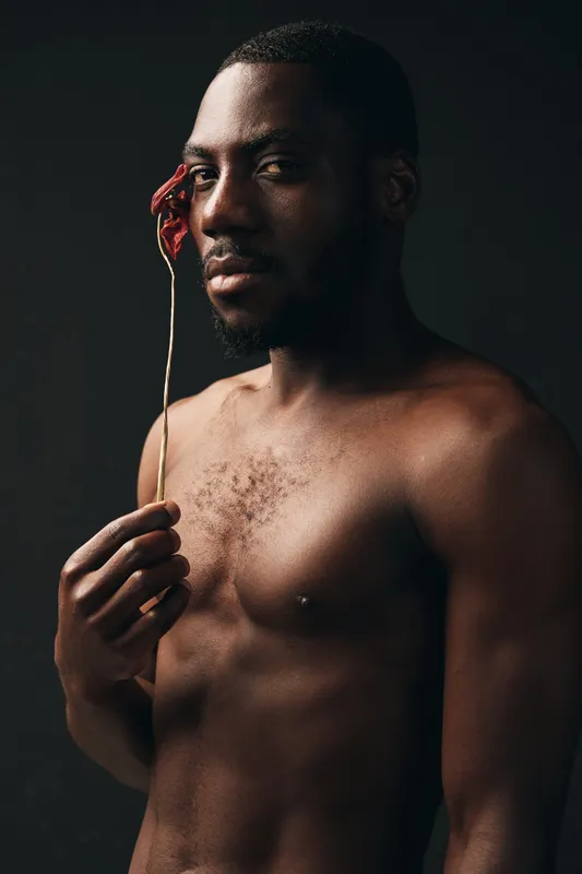 Creative conceptual portrait of African man with bare chest holding dried flower near face in dark studio. Artistic visual style with focus on skin texture and moody lighting for emotional impact.