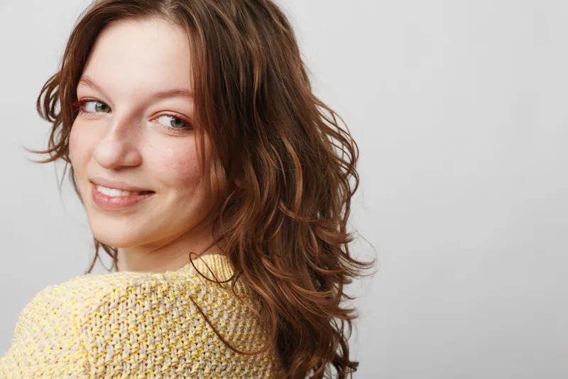 Smiling young woman with wavy brown hair wearing yellow knitted sweater looks over shoulder on light background. Concept of happiness, casual style, natural beauty, and positive mood.
