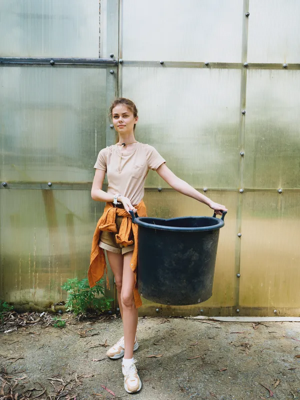 Woman bucket greenhouse gardening girl gardener container plants young female in casual jacket and sneakers holding large black bucket by polycarbonate wall, portrait of outdoor gardening activity.