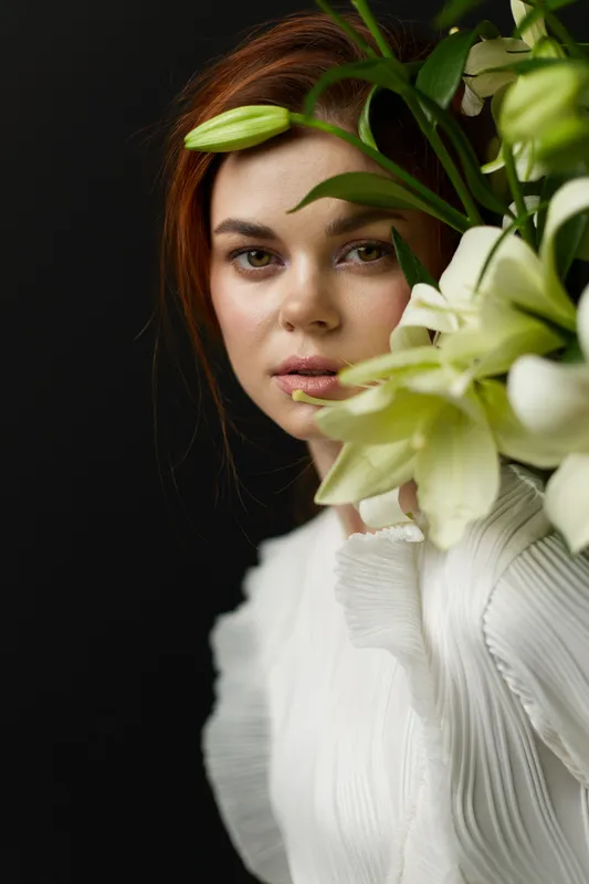 Portrait of a serene young woman with long brown hair, adorned with white lilies, wearing a textured white blouse, set against a dark backdrop, embodying beauty and tranquility