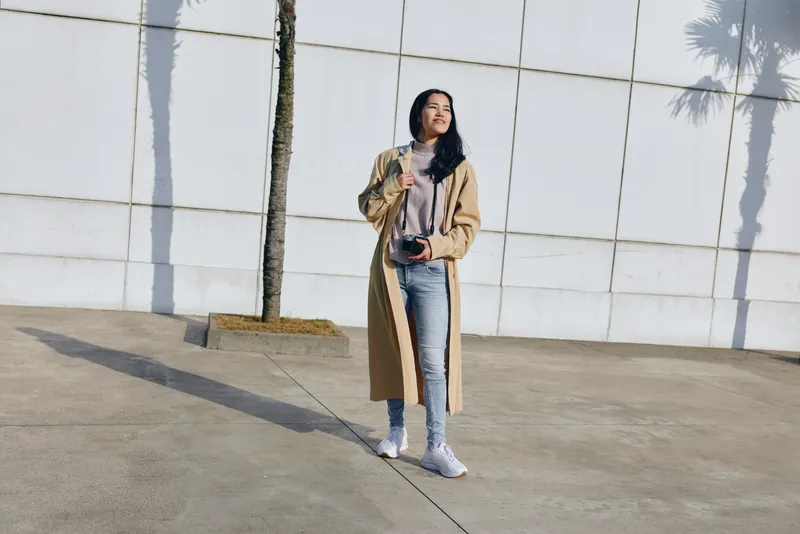 woman, camera, outdoor, coat, fashion, casual captured as a stylish photo session in bright daylight beside a pale wall and palm shade, presenting a relaxed street fashion moment