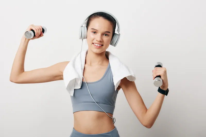 Young woman fitness portrait holding dumbbells with towel draped over shoulders, wearing headphones and smartwatch, smiling in grey activewear against clean white studio background.
