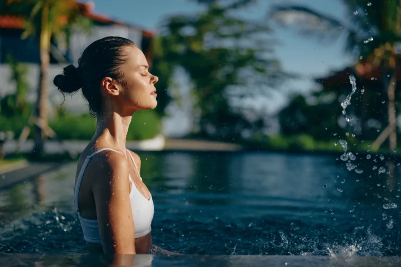 Young woman in white swimsuit relaxing in outdoor pool with eyes closed enjoying water splash and sunlight. Lifestyle concept of leisure, wellness, and summer vacation.