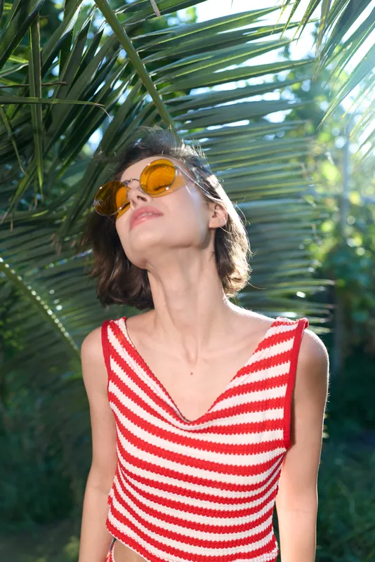 A stylish young woman wearing a red and white striped top stands outdoors beneath palm leaves, enjoying sunny weather. She wears round sunglasses and tilts her head slightly, radiating carefree