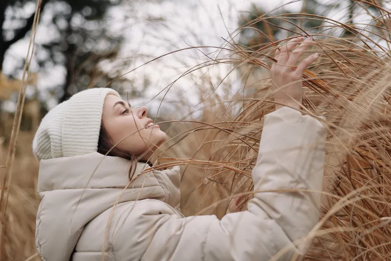 Woman in winter coat reaching through tall grasses in a quiet outdoor setting, capturing a peaceful, contemplative moment amid nature and grassy field with soft natural light.
