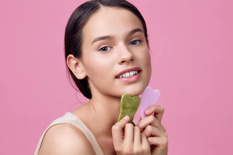 Young woman with jade and rose quartz gua sha tools against a pink background, promoting beauty and skincare routines for glowing skin