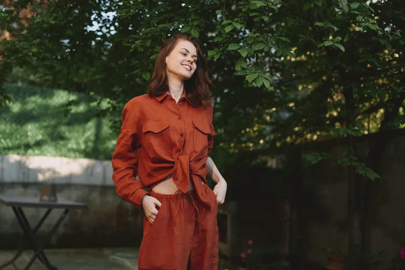 Smiling woman with wavy hair wearing a rust shirt and matching pants outdoors. Casual lifestyle fashion, confidence, and warmth under natural daylight. Lush green background, relaxed pose, everyday