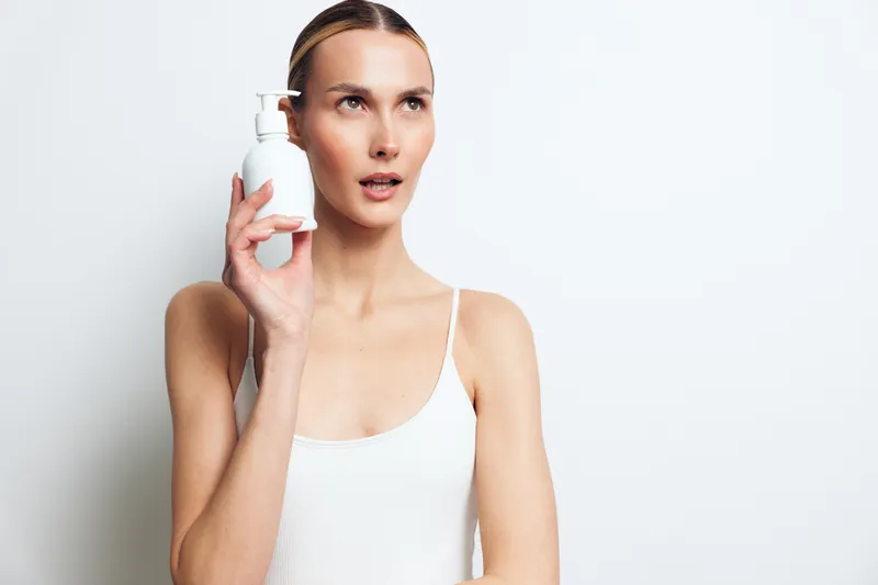 Young woman with clear skin holding white cosmetic bottle near face, wearing white tank top, studio portrait with plain background, skincare concept, beauty product