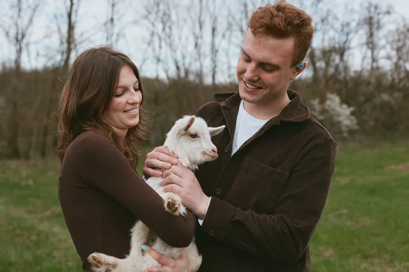 Happy couple outdoors with a goat kid, sharing affection and smiles, as they hold the young animal in a green countryside setting, capturing a joyful bonding moment and warm family atmosphere.