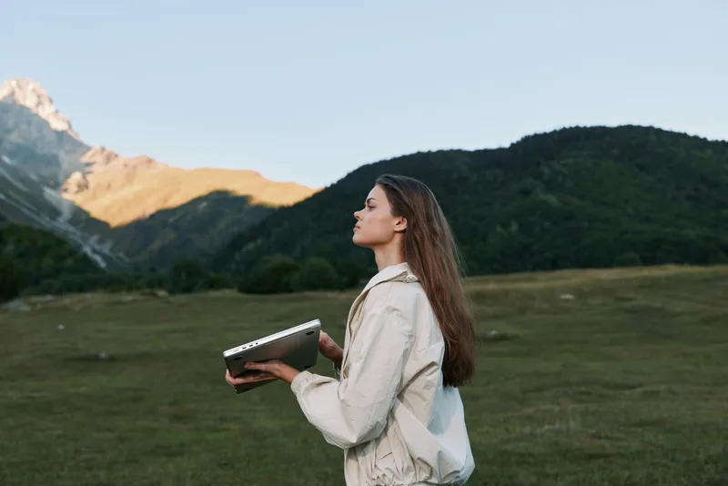 A thoughtful young woman stands in a vast meadow, holding a notebook as the sun casts warm light on distant peaks, conveying focus, curiosity, and creative outdoor work.
