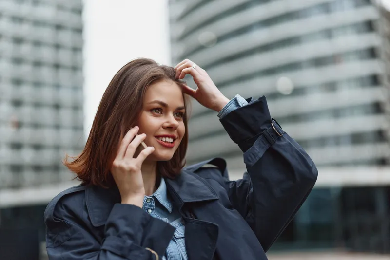 Young woman with brunette hair smiles while talking on a cellphone on a city street, wearing a denim shirt under a dark jacket, with glass buildings in the background.