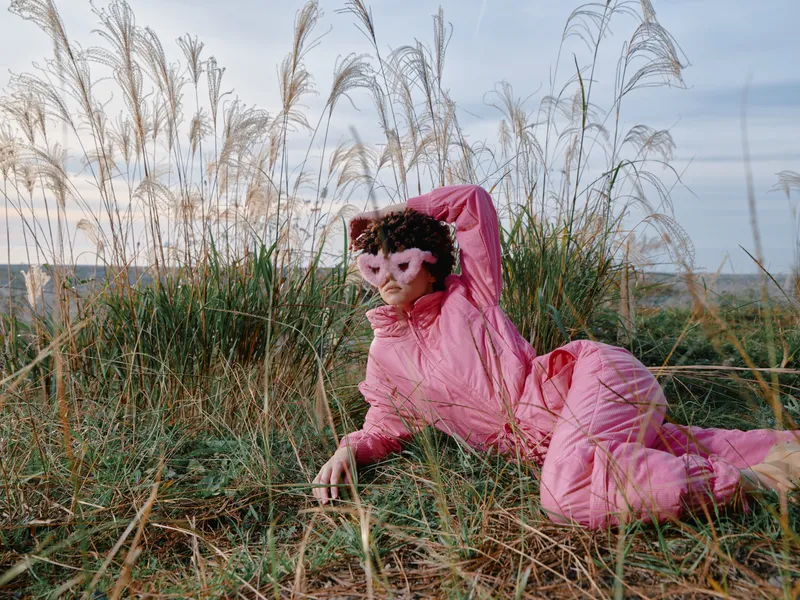 Person in pink costume lying in outdoor grass near a beach, wearing a monkey mask and relaxed pose, playful surreal scene with shore breeze and soft daylight.