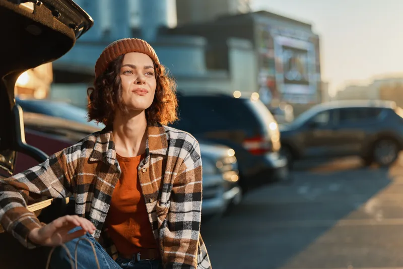 Woman portrait candid golden hour urban car lifestyle authenticity, young woman sitting by open trunk in parking lot with warm golden hour glow, mindful living and emotional storytelling