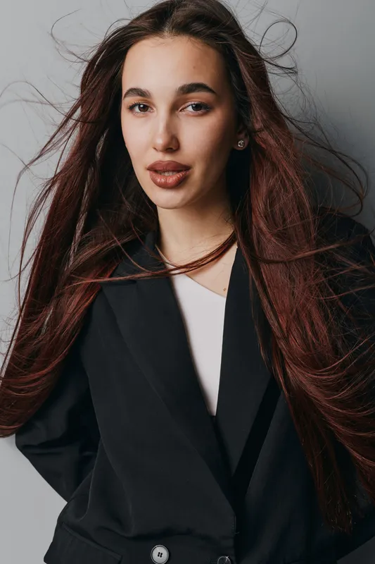 Woman with long hair and natural makeup wearing black blazer posing in studio. Portrait of confident female model with flowing hair and neutral expression on gray background.