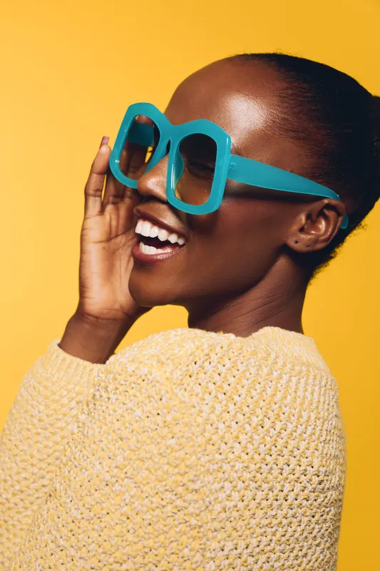 Happy African woman with dark skin wearing large blue sunglasses and yellow sweater smiles brightly against yellow background. Concept of fashion, joy, and summer style.