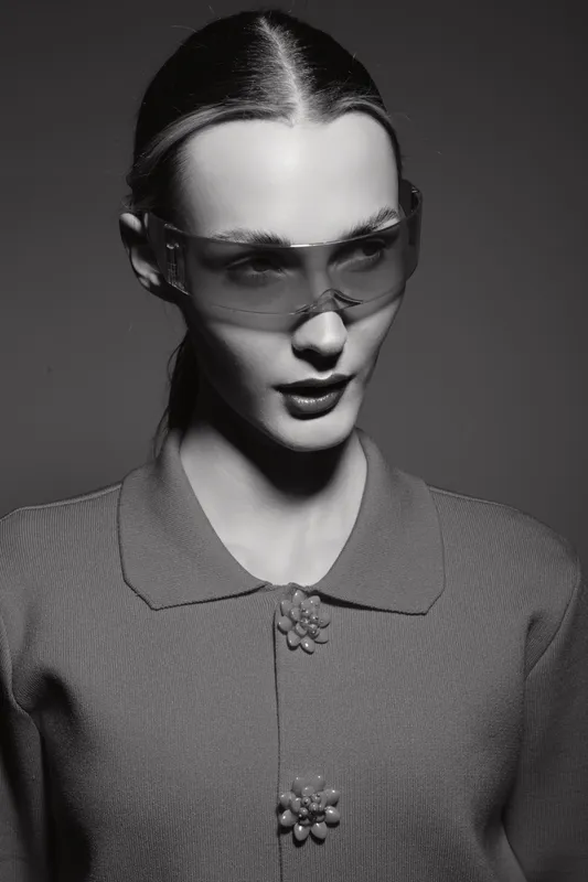 Portrait of a slim white woman with pale skin wearing transparent protective glasses and a textured buttoned top. Studio lighting creates high contrast and sharp shadows. Fashion, eyewear.