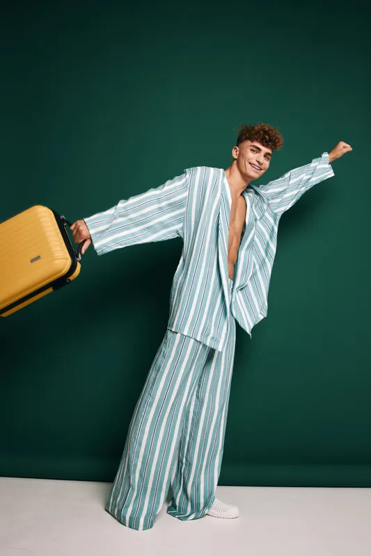 Young man with curly hair wearing striped oversized suit holds yellow suitcase with wheels posing standing on white floor against dark green studio background with confident smile.