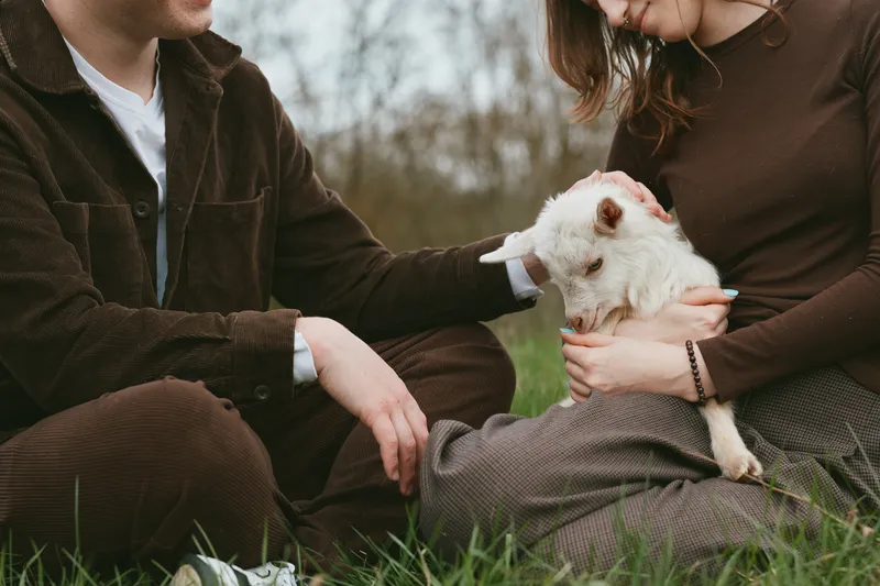 A couple sits on grass, gently holding a small goat kid between them, sharing a quiet moment outdoors, with soft natural light and a warm, intimate mood focused on care and companionship.