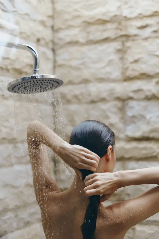 Woman taking outdoor shower with natural stone wall background, wet skin, and long dark hair tied back in a peaceful and refreshing moment.
