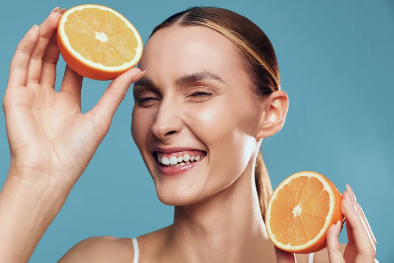 Young woman, orange citrus, smile on blue background, holds fresh orange halves near face, joyful expression, vitamin concept, healthy lifestyle, skincare for beauty