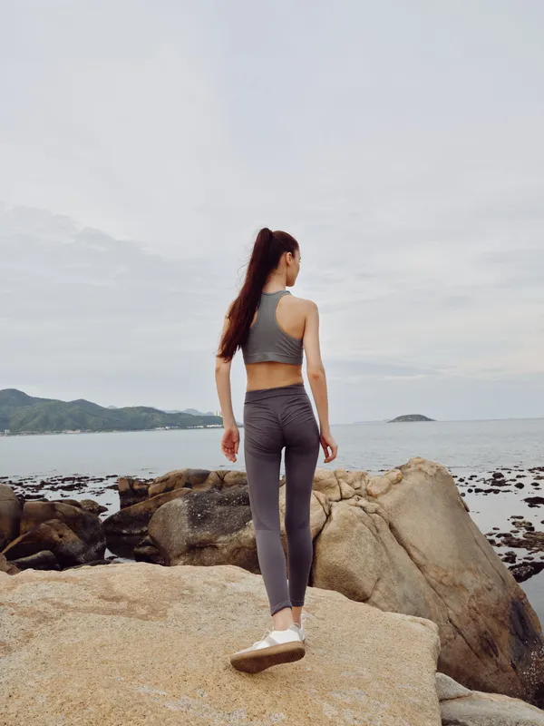 Young model in athletic wear standing on rocky beach, promoting fitness and wellness with serene ocean backdrop and natural colors