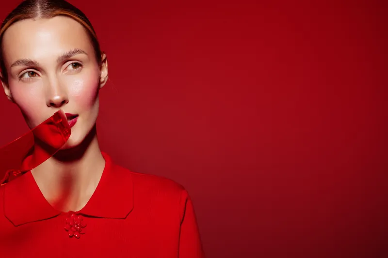 Young woman in red sweater with red ribbon on lips looks away on red background. Concept of fashion, beauty, and creative portrait with vibrant color and minimalistic style.