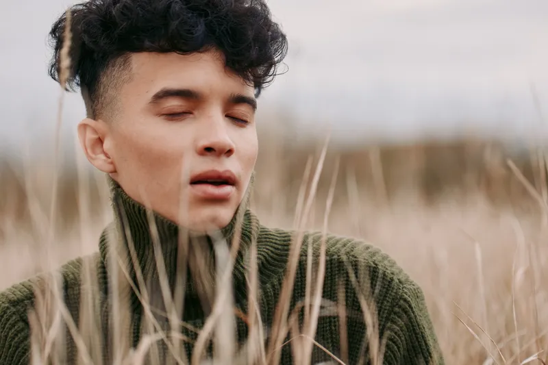 A candid close portrait of a young person with closed eyes in tall grasses, conveying authenticity and genuine emotional presence, tactile sweater texture, natural light and unposed expression.