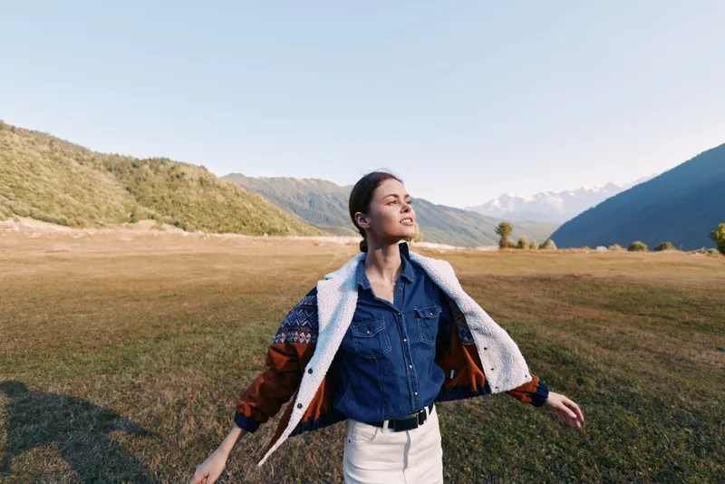 Woman in nature on a meadow with mountains in the background, wearing a jacket, arms open embracing freedom and travel outdoors, smiling and enjoying a scenic peaceful landscape.
