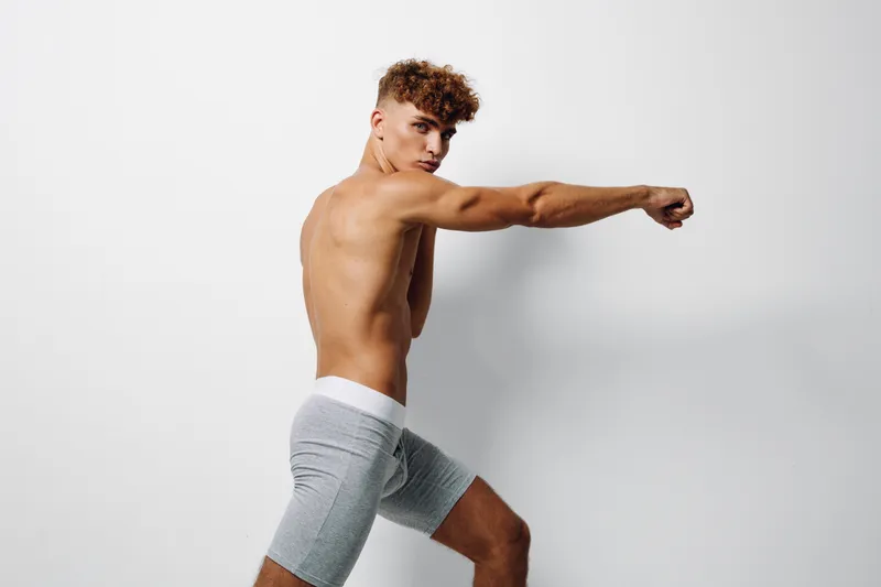 Young man with curly hair in gray shorts posing shirtless against white background, showing athletic build and focused expression in studio lighting.