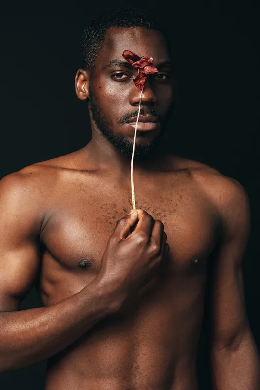 Creative conceptual portrait of African man with bare chest holding dried flower in front of face against dark background. Artistic visual style with focus on expression and texture.