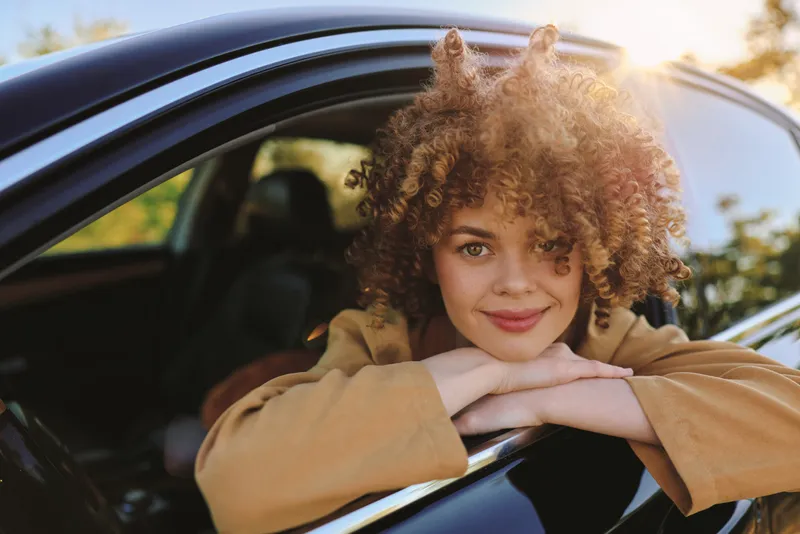 Outdoor portrait of a woman leaning on a car window, smiling softly, sunlit glow, casual fashion, natural tones, curly hair, relaxed mood, retro film color vibe, gentle breeze