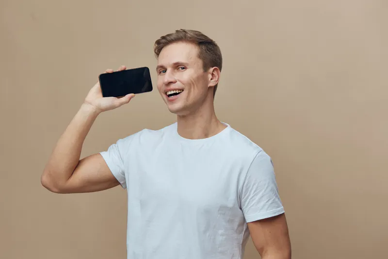 Smiling young man taking a selfie with smartphone, casual white t-shirt, cheerful expression, brown hair, light background, lifestyle concept, modern technology, leisure activity, friends, social medi