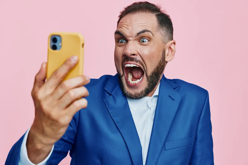A professional business man captures a dynamic selfie on a solid pink backdrop, showing vivid emotion and energy as he engages with a phone.