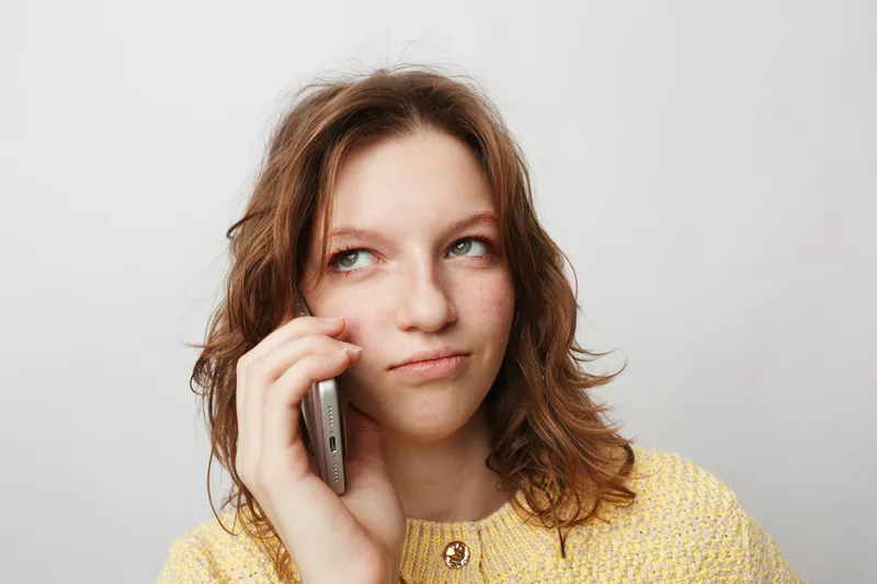 Young woman with curly hair in yellow sweater talks on smartphone, looking thoughtful and slightly skeptical. Isolated on white background, concept of communication and emotion.