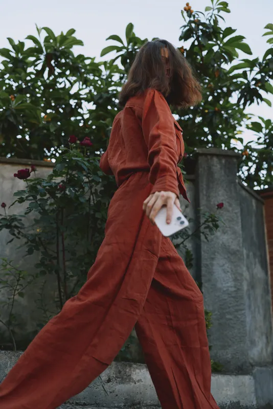 Vital woman in rust jumpsuit walks outdoors holding phone, motion captured in an urban garden setting, lifestyle portrait emphasizing movement and contemporary fashion