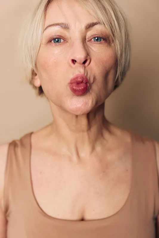 Mature woman with light hair makes a playful kiss face close up. Nude tone top and soft beige background create a natural and warm portrait concept of beauty and expression.