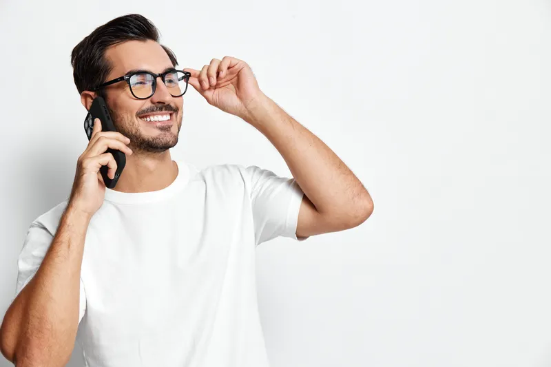 Smiling man wearing glasses talking on mobile phone, looking cheerful and happy against plain solid white background. People lifestyle concept.