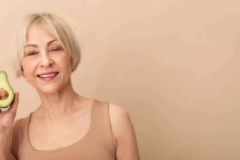 Smiling mature woman holds half of avocado, wearing beige tank top, isolated on beige background. Concept for healthy lifestyle, nutrition, and natural wellness for seniors.