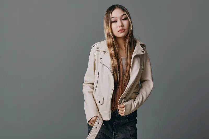 Young woman with long straight hair wearing beige jacket and casual clothes standing against gray background looking down with calm expression in studio portrait.